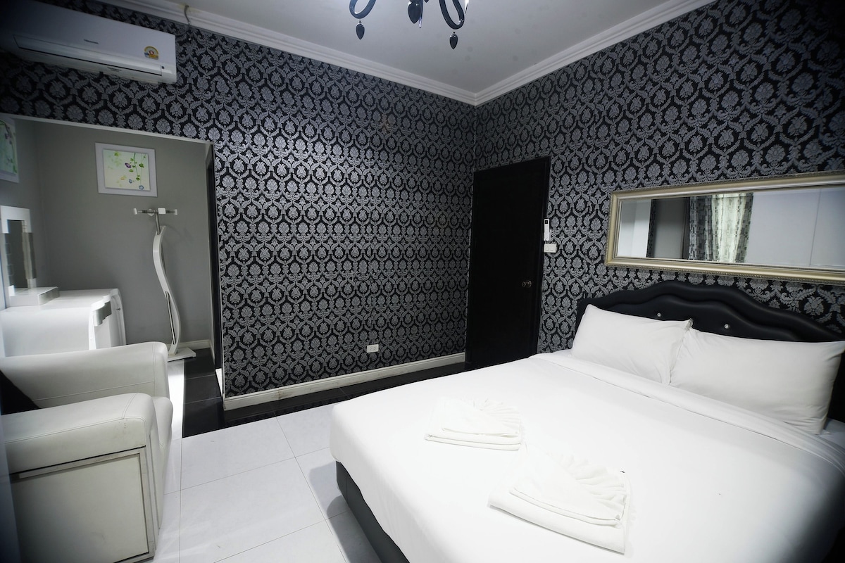 A hotel room with a patterned wallpaper, a bed with white linens, a bedside table, a mirror, and a shower area.