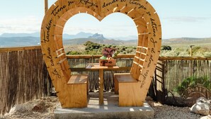 A wooden heart-shaped structure with benches and a table, surrounded by a bamboo fence, overlooking a mountainous landscape.