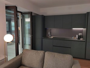 A modern kitchen with dark cabinets, a countertop, and a built-in oven. There is a sofa with a round light in front of a glass door leading to a balcony. - NJ Hotels & Apartments (Milan)