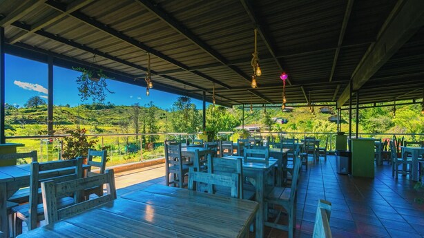 An outdoor dining area with wooden tables and chairs, a metal roof, and a view of a green landscape. - Bamba Hotel Guatapa (Guatapé)