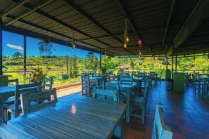 Un espacio al aire libre para comer con mesas y sillas de madera, techo de metal y vistas a un paisaje verde.