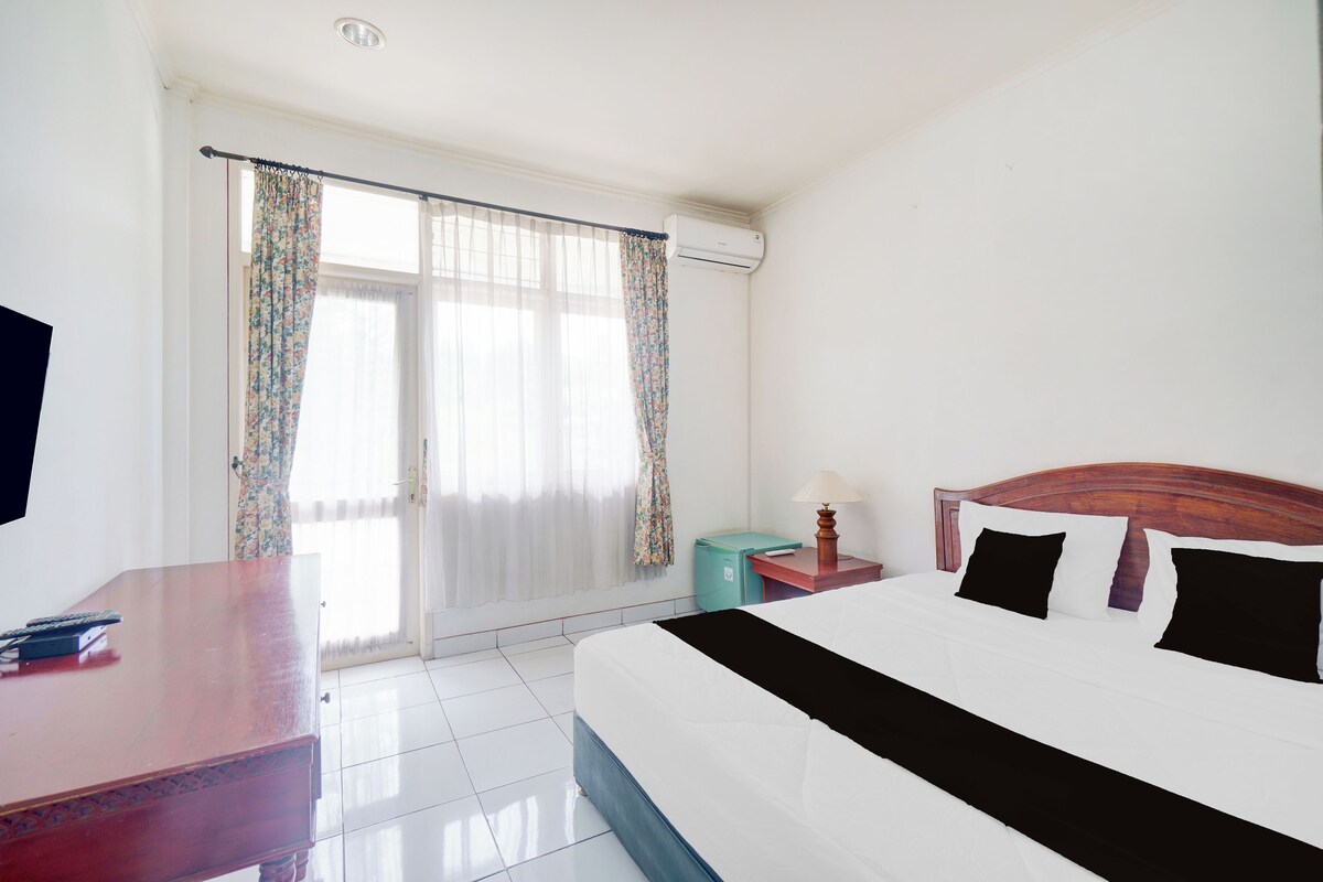 Habitación de hotel con cama, escritorio, silla, lámpara, televisor y ventana con cortinas.
