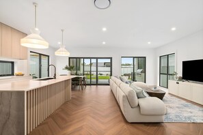 A modern kitchen with a built-in island, a dining area with a table and chairs, a living room with a sofa and a TV, and large windows offering a view of the outdoors. - The Dunes on Beecroft (Huskisson)