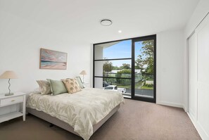 A bedroom with a bed, nightstand, lamp, painting, and a large window with a view of the outdoors. - The Dunes on Beecroft (Huskisson)