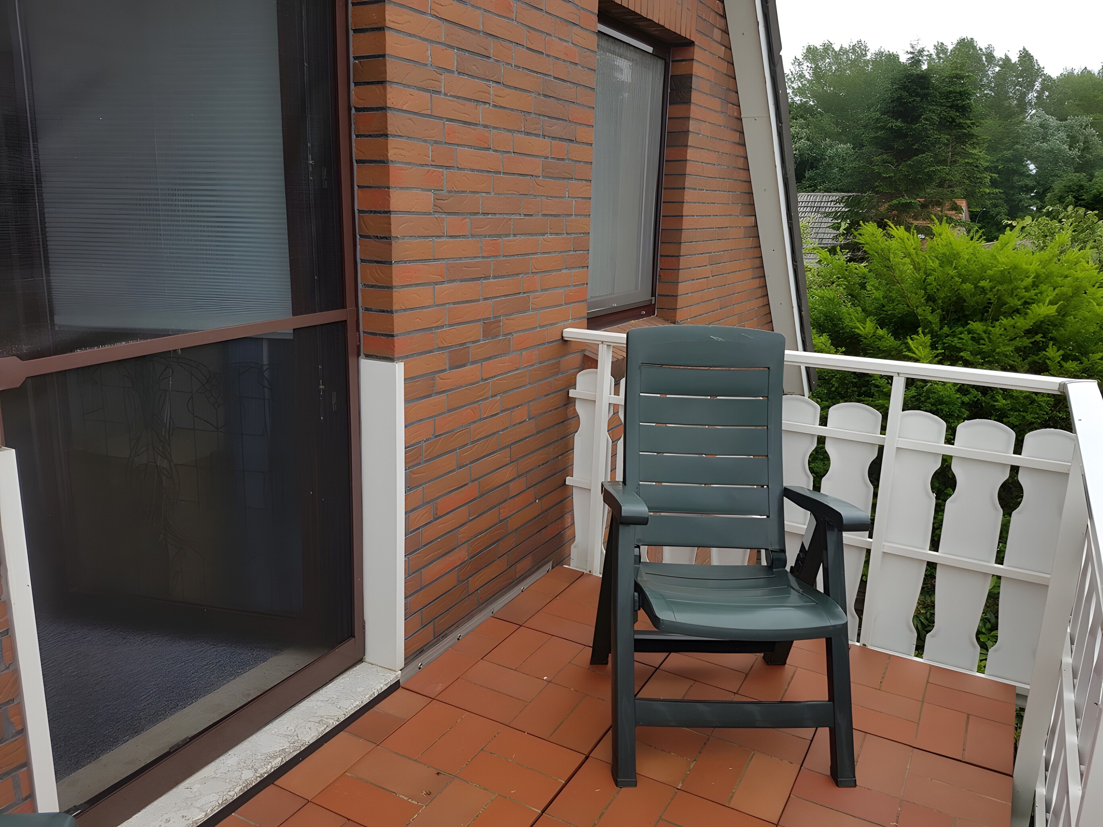Ein Balkon mit einem Stuhl, ein Fenster mit Rollläden und ein weißer Zaun.