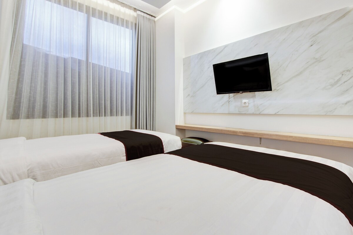Habitación de hotel con dos camas, un televisor montado en una pared de mármol y una ventana con cortinas.