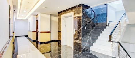 A modern hotel lobby with a marble floor, a staircase with glass railings, and a view of the sky through the windows.