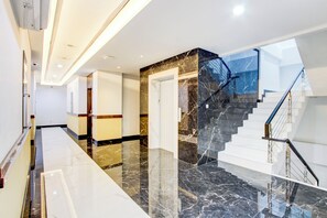 Um saguão de hotel moderno com piso de mármore, escada com corrimão de vidro e vista do céu pelas janelas.