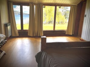 Makuuhuoneessa on suuri sänky, puulattia, ikkuna, jossa on verhot, ja ikkunasta avautuu näkymä järvelle ja puille.