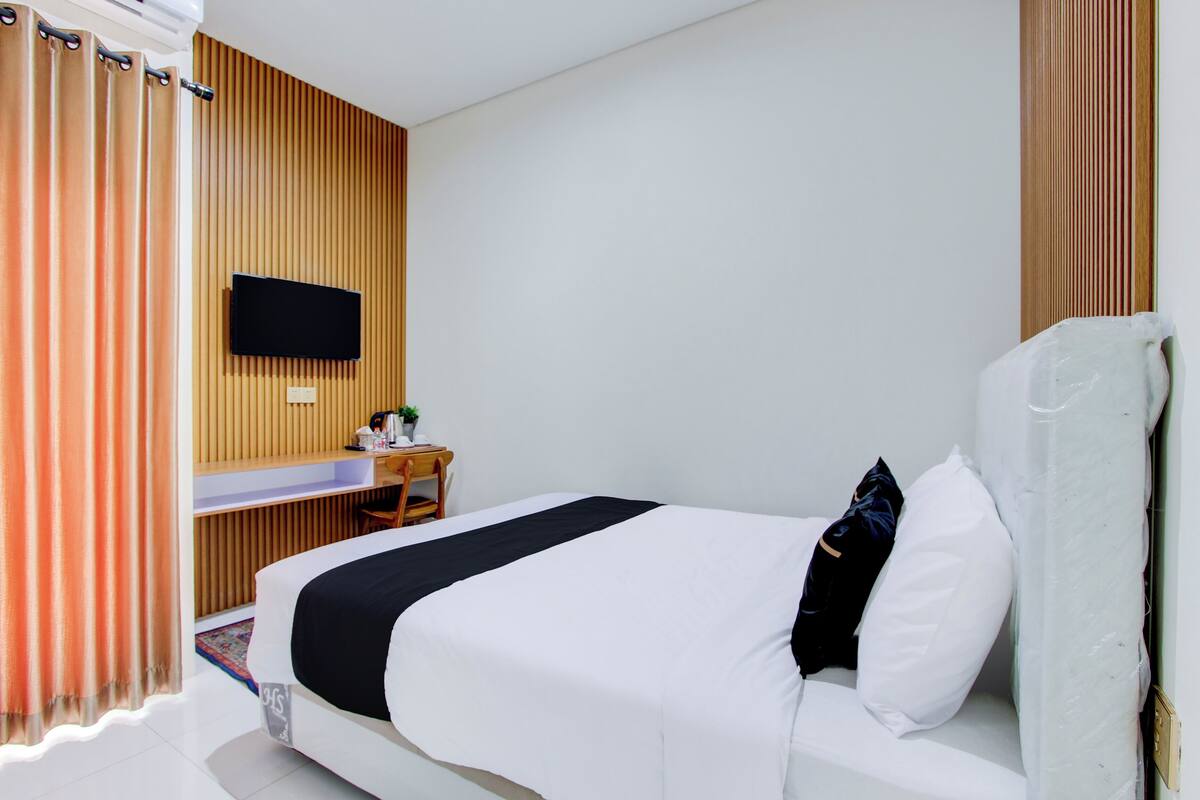 A hotel room with a bed, a desk, a chair, a TV mounted on the wall, and a window with curtains.