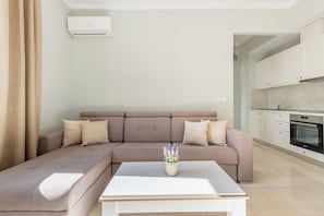 Ein modernes Wohnzimmer mit einem Ecksofa, einem weißen Couchtisch und einer Küchenzeile mit weißen Schränken und einem eingebauten Backofen.