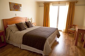 Ein Schlafzimmer mit einem hölzernen Bett, einem Nachttisch mit Lampe, einem Stuhl, einem Tisch und Blick ins Freie durch ein Fenster mit Vorhängen.