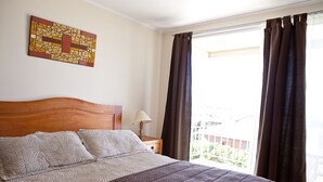 A bedroom with a bed, a nightstand, a lamp, a window with curtains, and a wall art piece. - Hotel Y Cabañas Portal Del Inca (Caldera)