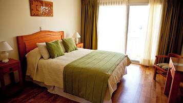 A bedroom with a wooden bed, green pillows, a bedside table with a lamp, a chair, and a window with curtains.