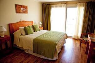 Un dormitorio con cama de madera, almohadas verdes, una mesita de noche con lámpara, una silla y una ventana con cortinas.