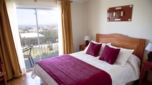 Una camera d'albergo con un letto grande, un balcone vista, una testiera in legno e un comodino con una lampada.