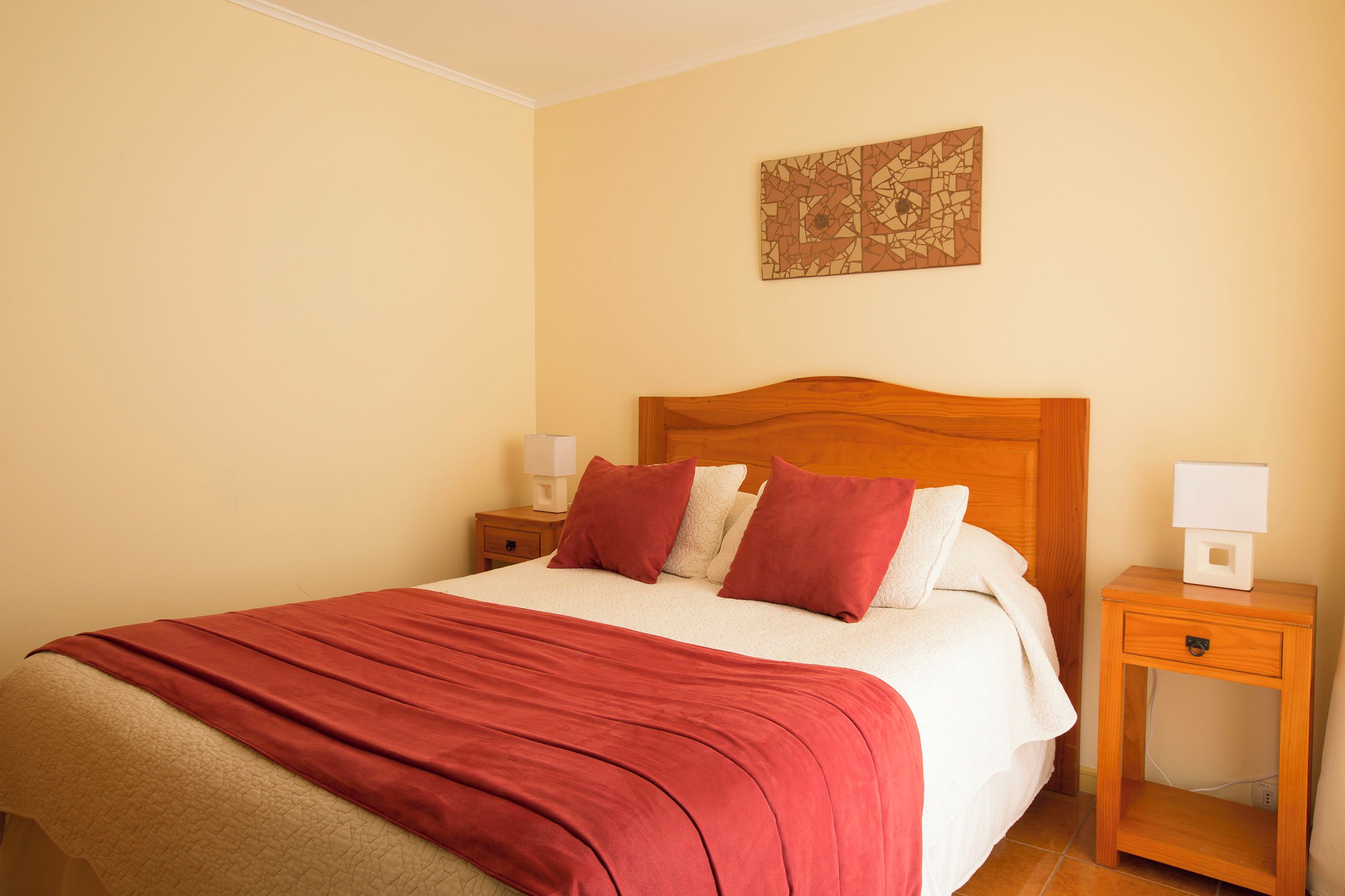 A neatly made bed with a red blanket, a wooden headboard, and a bedside table with a lamp.