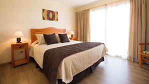 Un letto rifatto con cura, una testiera in legno, due comodini con lampade, un quadro appeso al muro e vista sull'esterno attraverso ampie finestre.