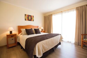 A neatly made bed with a wooden headboard, two bedside tables with lamps, a wall art piece, and a view of the outdoors through large windows. - Hotel Y Cabañas Portal Del Inca (Caldera)