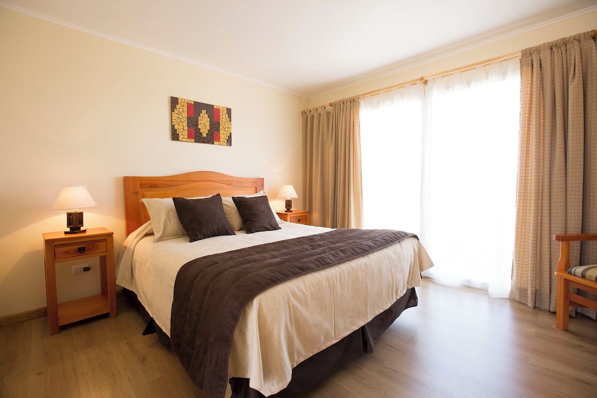 A neatly made bed with a wooden headboard, two bedside tables with lamps, a wall art piece, and a view of the outdoors through large windows.