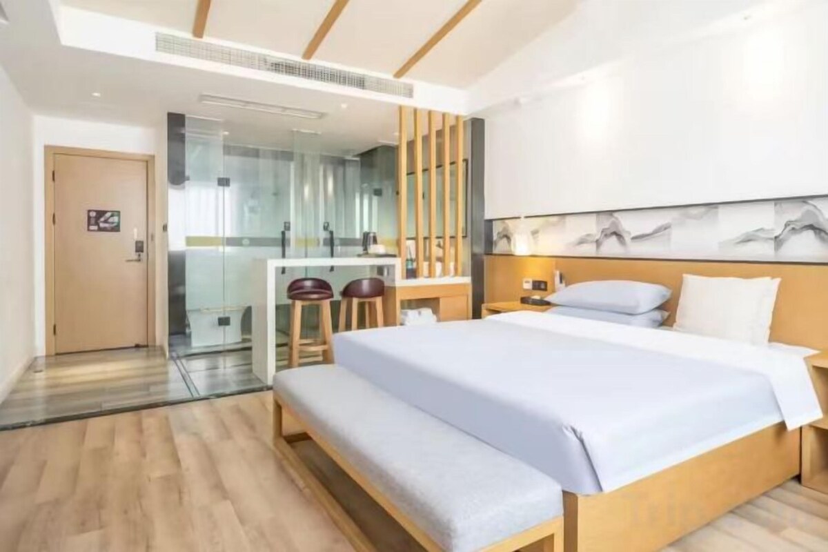 A modern hotel room with a large bed, a desk with a chair, and a glass partition.