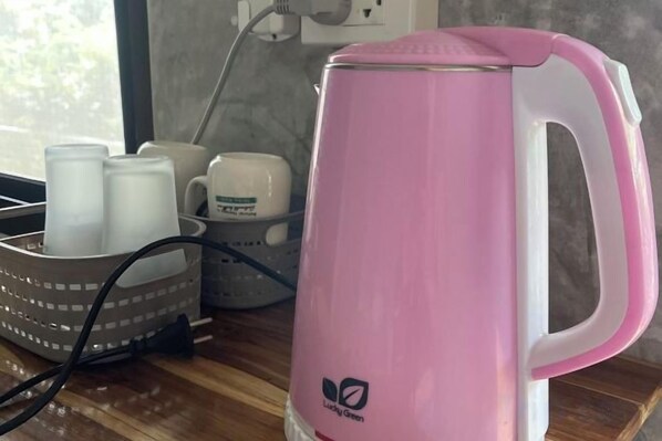 Una tetera eléctrica rosa sobre una superficie de madera, junto a una cesta de tazas blancas y un enchufe al fondo.