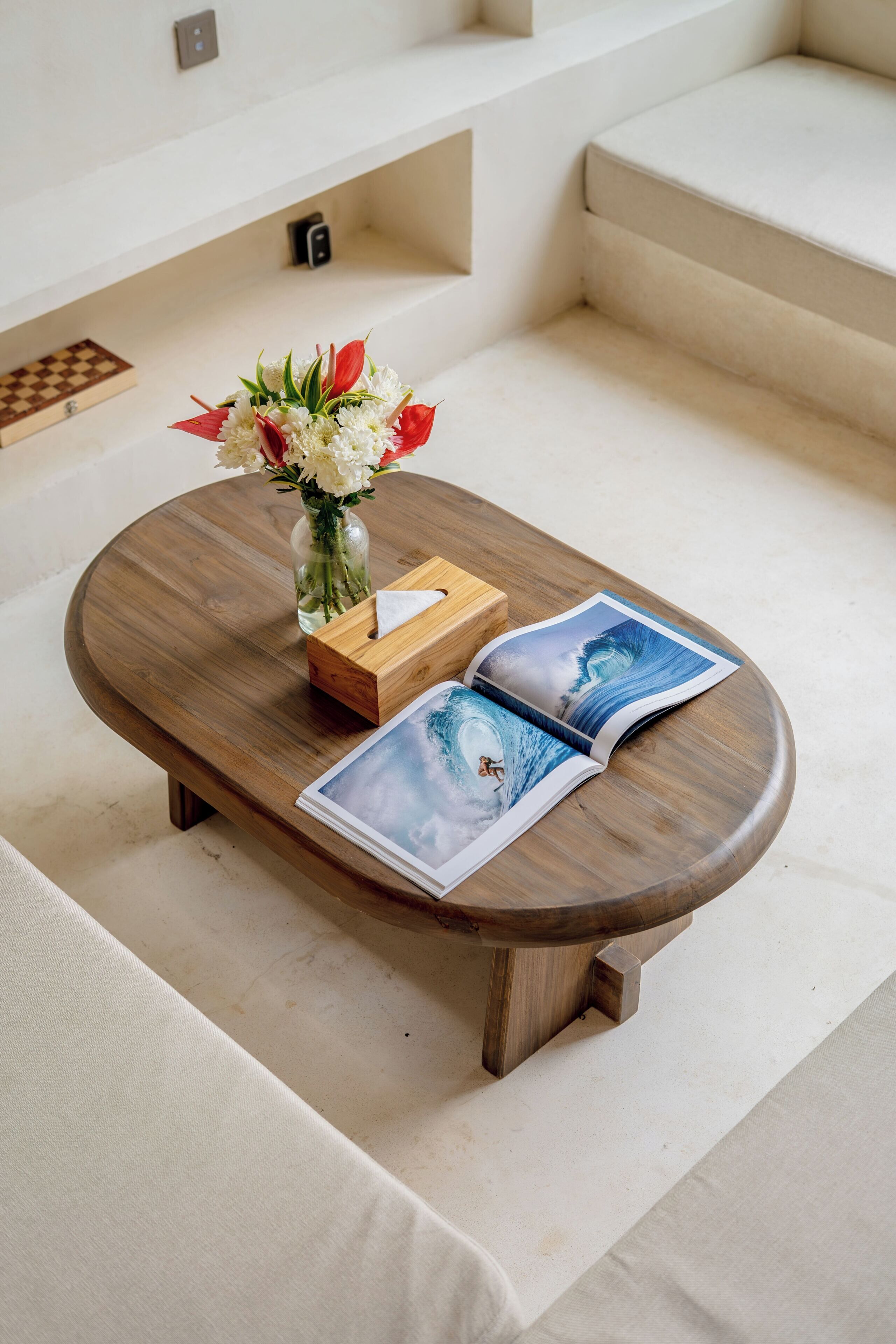 A wooden coffee table with a vase of flowers and two magazines on a white carpeted floor.