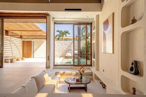 A modern interior with a sofa, coffee table, and a view of a pool and palm trees. - Aozora Villas Uluwatu (Uluwatu)