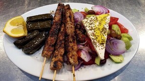 Un plato con brochetas de carne a la parrilla, hojas de parra rellenas y una ensalada de tomates, pepinos y aceitunas.