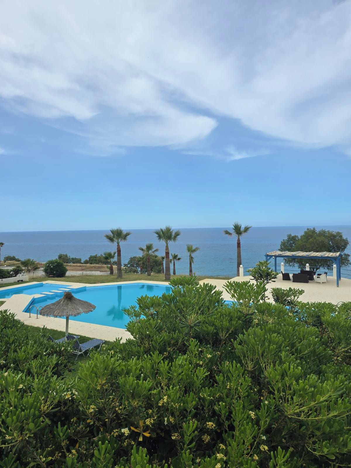 Una piscina con vistas al mar, rodeada de vegetación y una zona de estar con sombra.