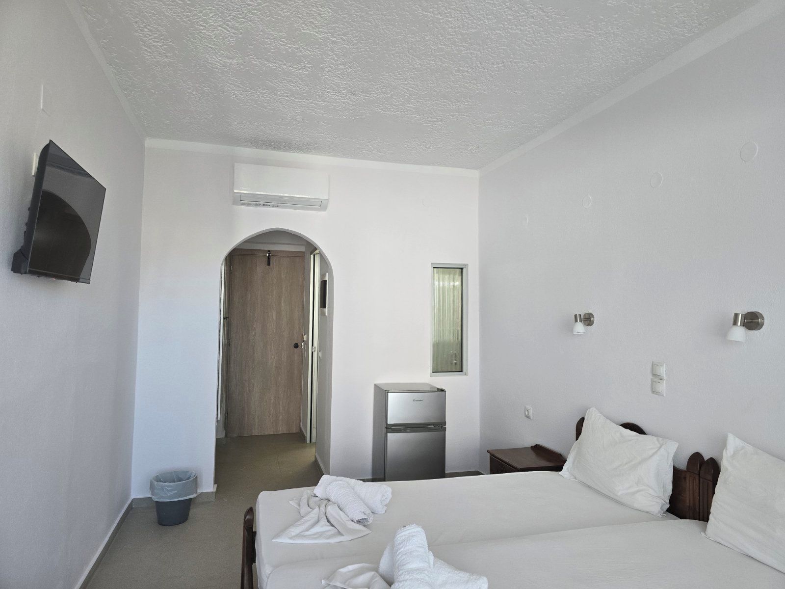 Une chambre d’hôtel avec un lit, un petit bureau, un miroir, une télévision et une porte donnant sur un couloir.