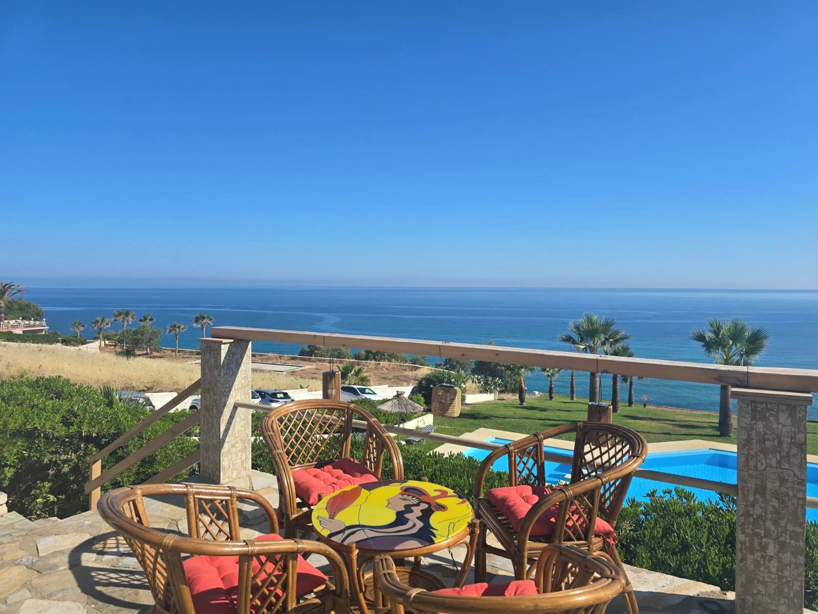 Una terraza con sillas de mimbre y una mesa, con vista a una piscina y el océano.