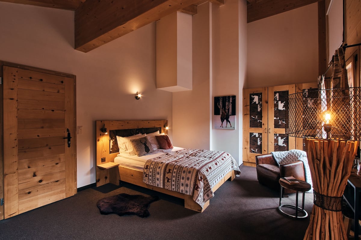 Dormitorio con estructura de madera, cama, mesitas de noche, una silla y una lámpara fijada a la pared.