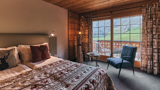A bedroom with a large bed, a chair, a small table, and a window with a view of a green landscape. - Etable Des Crosets (Val d'Illiez)