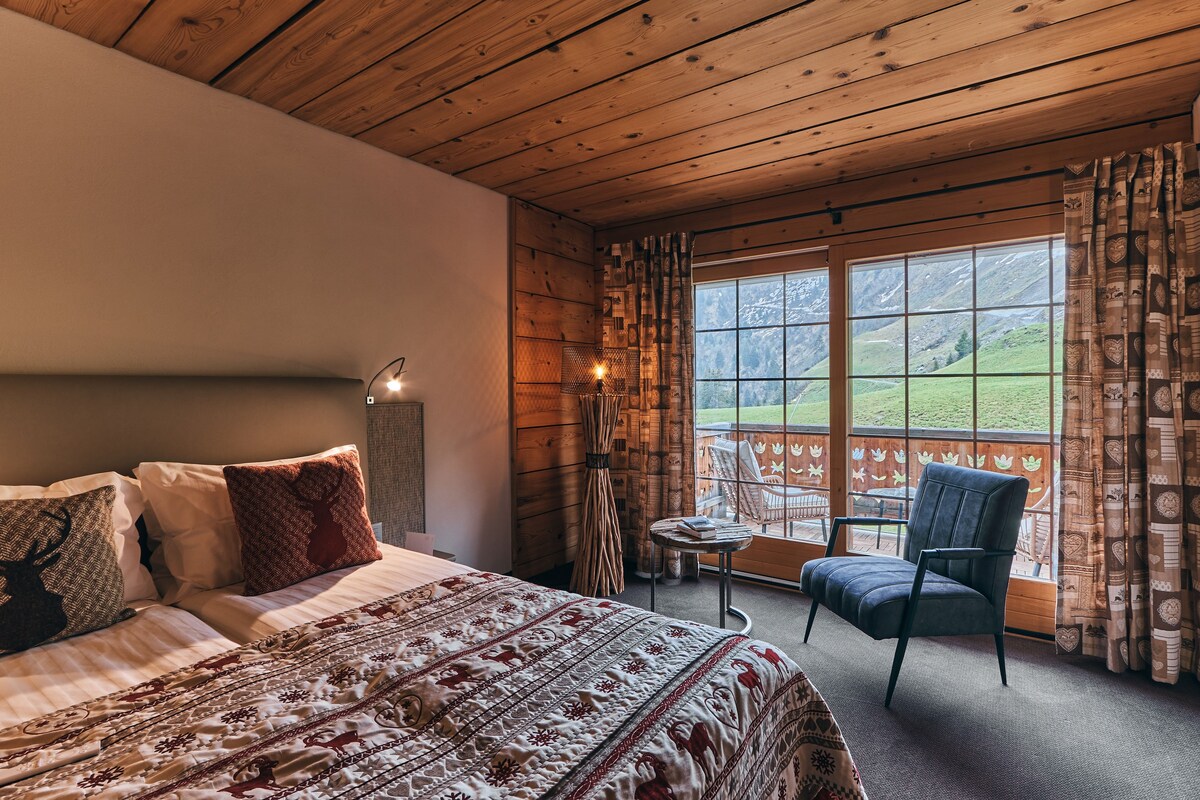 Un dormitorio con una cama grande, una silla, una mesita y una ventana con vistas a un paisaje verde.