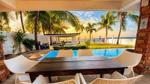 An outdoor pool area with a wooden table and white wicker chairs, surrounded by palm trees and a view of the ocean.