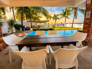 An outdoor pool area with a wooden table and white wicker chairs, surrounded by palm trees and a view of the ocean. - Brizo Beach Resort Bataan (Morong)
