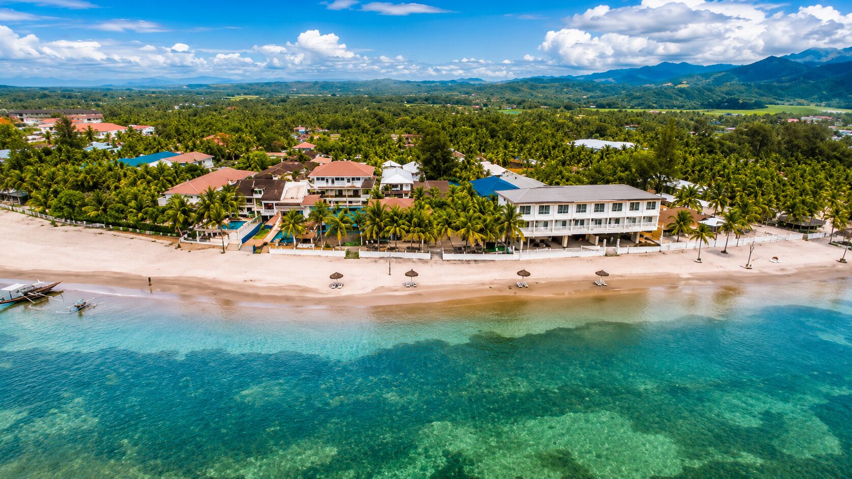 An aerial view of a coastal resort with a beach, clear waters, and lush greenery.