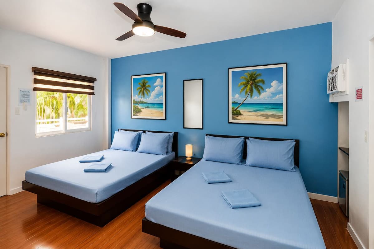 Quarto de hotel com duas camas, um ventilador de teto, uma janela com cortinas e duas quadros com imagens de praia na parede.