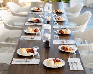 Uma longa mesa de jantar com cadeiras brancas, posta com pratos de comida, copos e talheres.