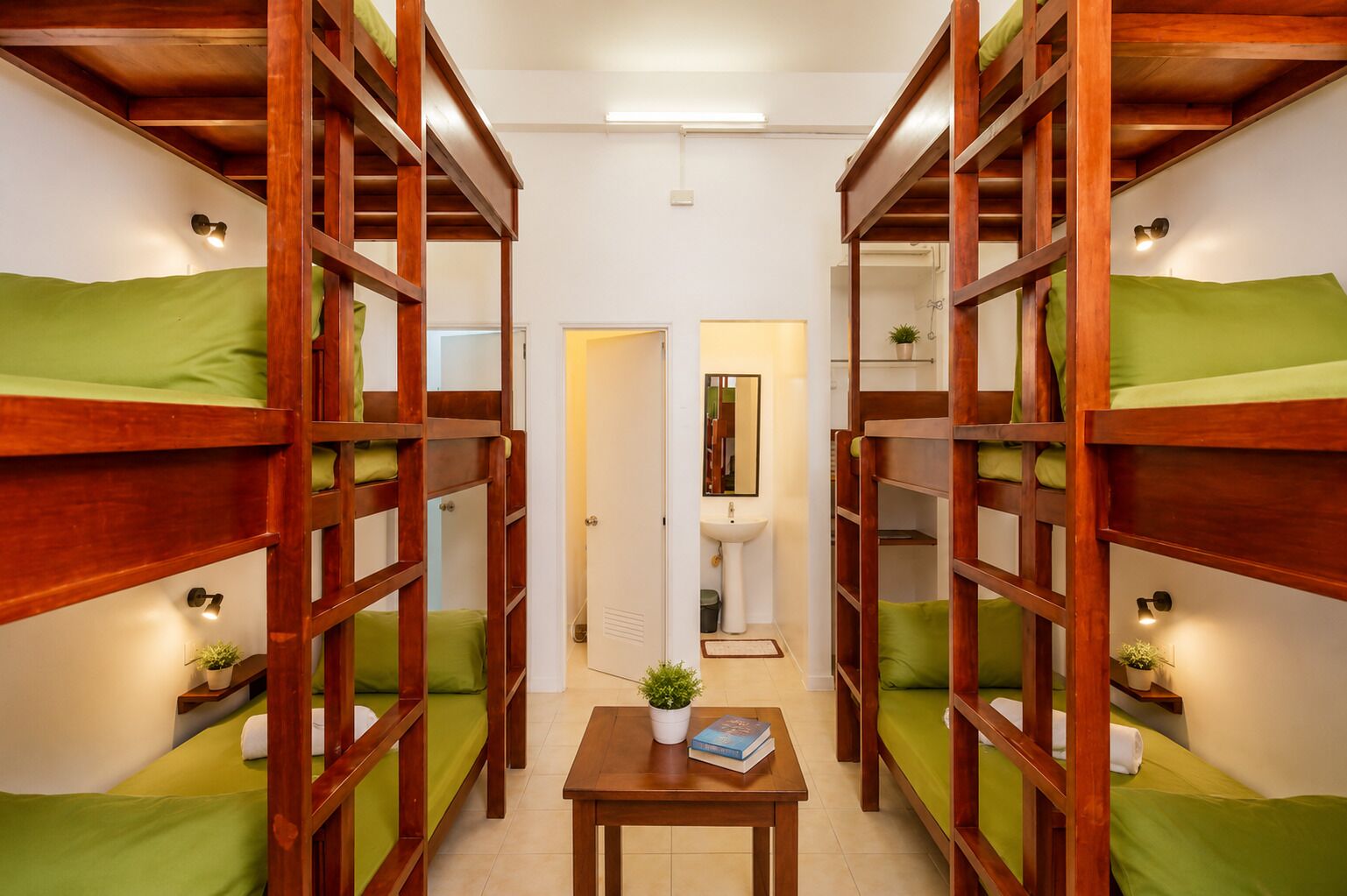 Una habitación con literas, una mesita con un libro y una planta, y un baño con lavamanos y espejo.