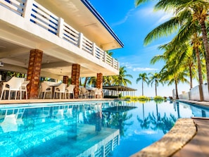 Zona junto a la piscina con vista despejada a la pileta, palmeras y un edificio de fachada blanca con detalles azules.