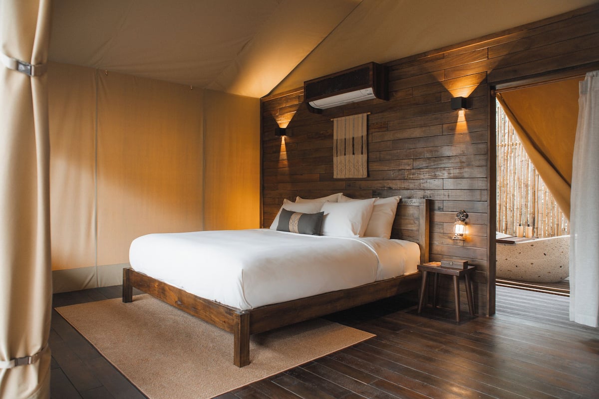 Ein Schlafzimmer mit einem Holzkopfteil, einem Bett mit weißer Bettwäsche, einem kleinen Nachttisch und einer Wand-Klimaanlage.