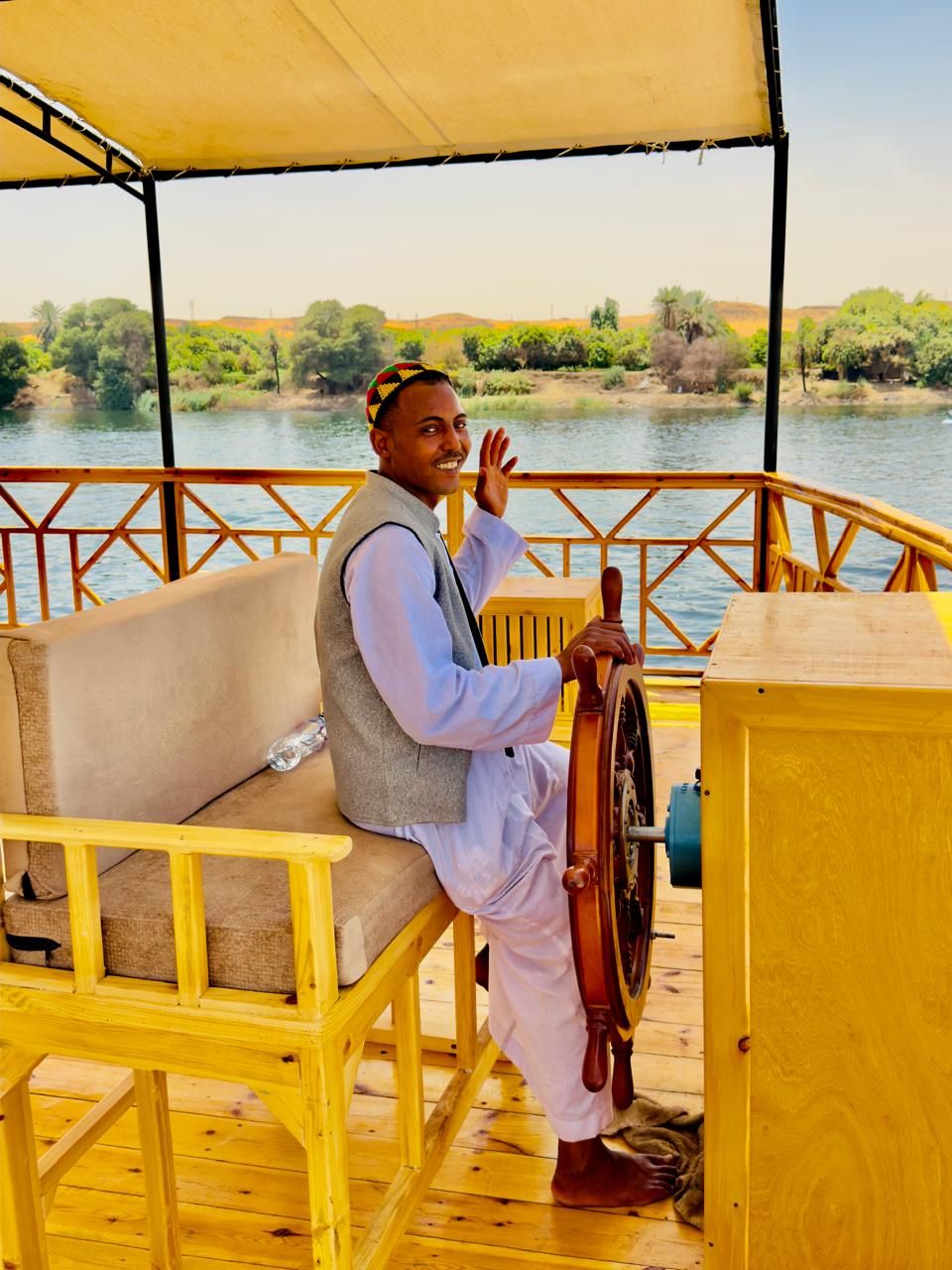 A person steering a boat on a wooden deck with a view of a river and greenery.