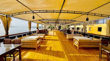 A boat interior with wooden flooring, tables, and benches, and a covered area with a view of the water.