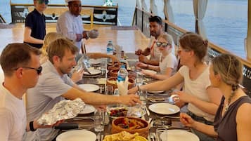 A group of people dining on a boat with a table set for a meal, surrounded by water and hills in the background.