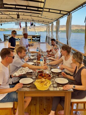 Un grupo de personas cenando en un barco, con una mesa preparada para la comida, rodeado de agua y colinas al fondo.
