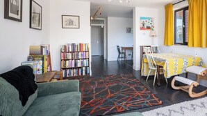 En stue med sofa, reol, spisebord med stole og et køkken i baggrunden.