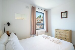Una camera d'albergo con un letto, una cassettiera, una lampada, una finestra con tende e un quadro appeso al muro.