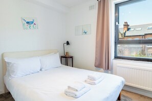 A hotel room with a bed, a nightstand, a lamp, and a window with a view of buildings. - Peaceful 2BDR Escape Near Primrose Hill (Greater London)